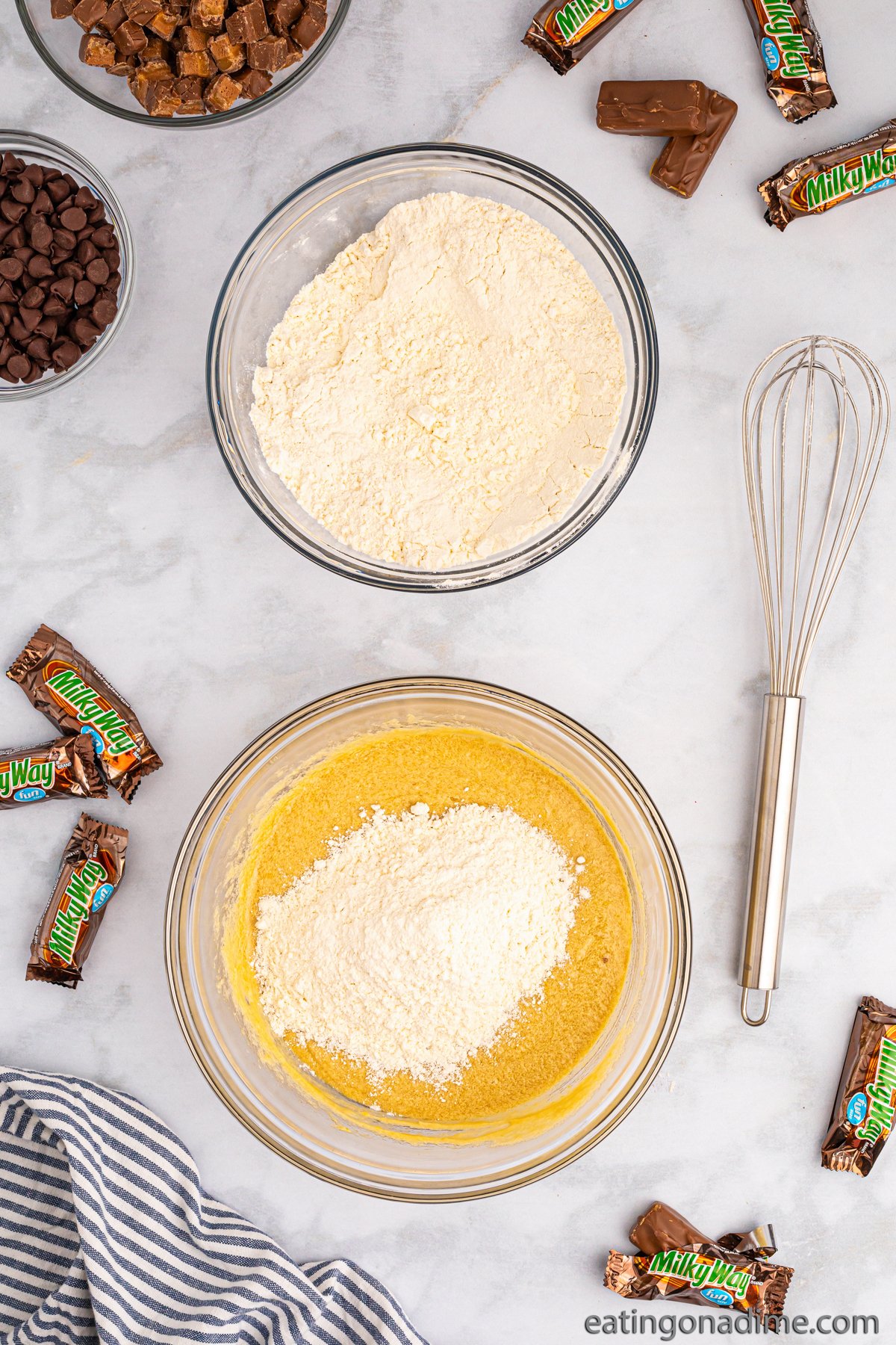 Combining the dry ingredients with the wet ingredients in a bowl with small bowls of chocolate chips and chopped Milky Way in bowls