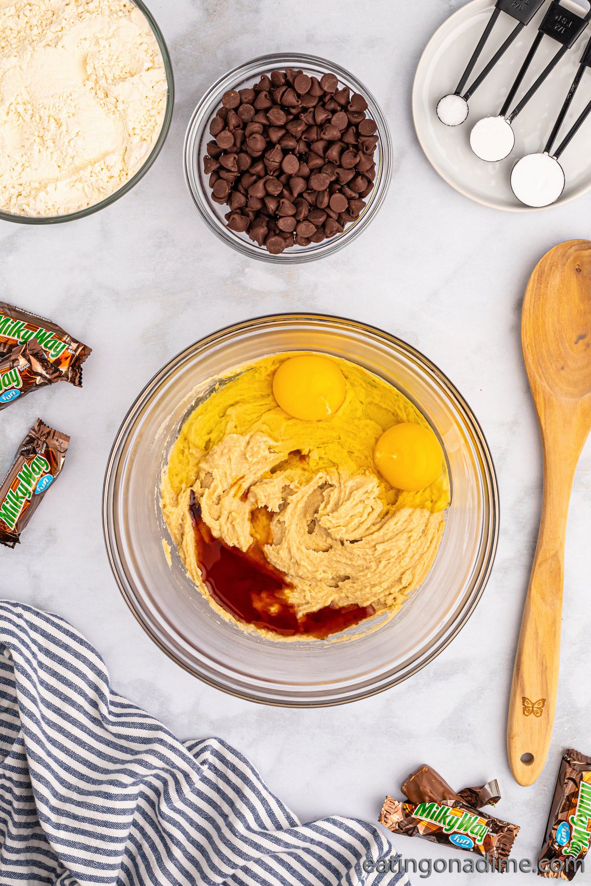 Mixing together butter, sugar, brown sugar and eggs in a clear bowl with a wooden spoon on the side. Small bowl of chocolate chips and flour on the side with teaspoons of dry ingredients