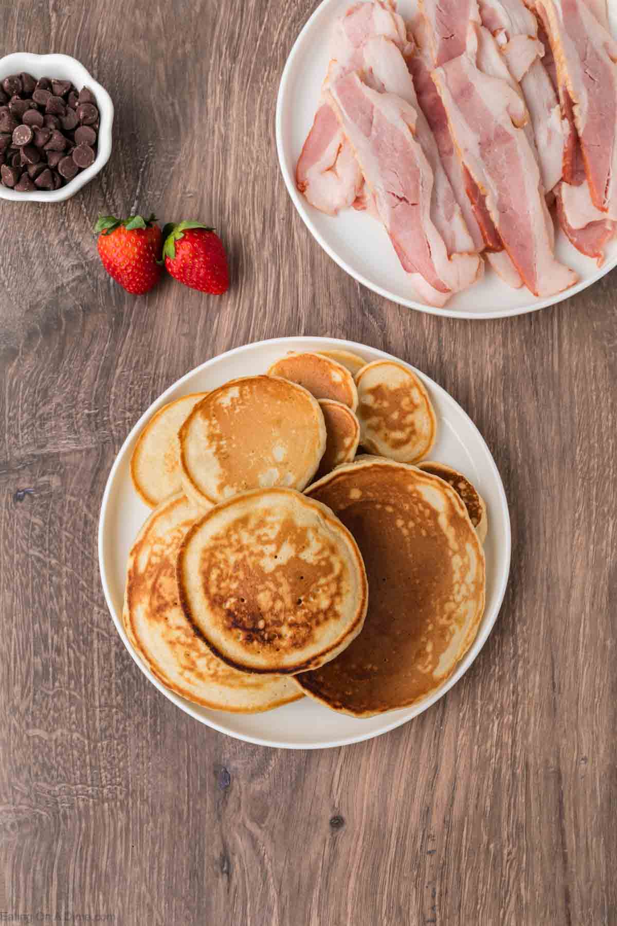 Cooked pancakes stacked on a plate with a plate of bacon on the side