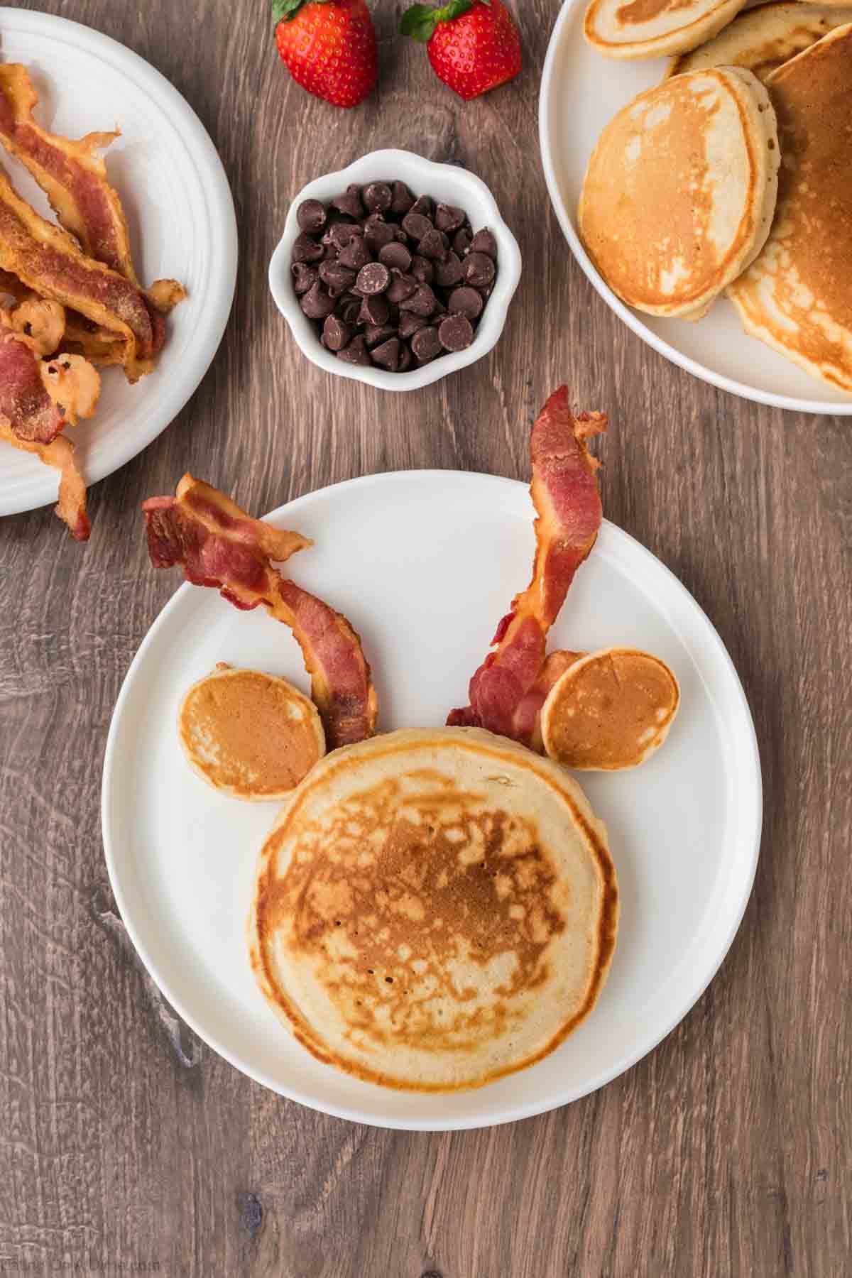 Pancakes formed into Reindeer face with the slices of bacon placed to make the horns on the reindeer with plates of bacon and pancake on the side