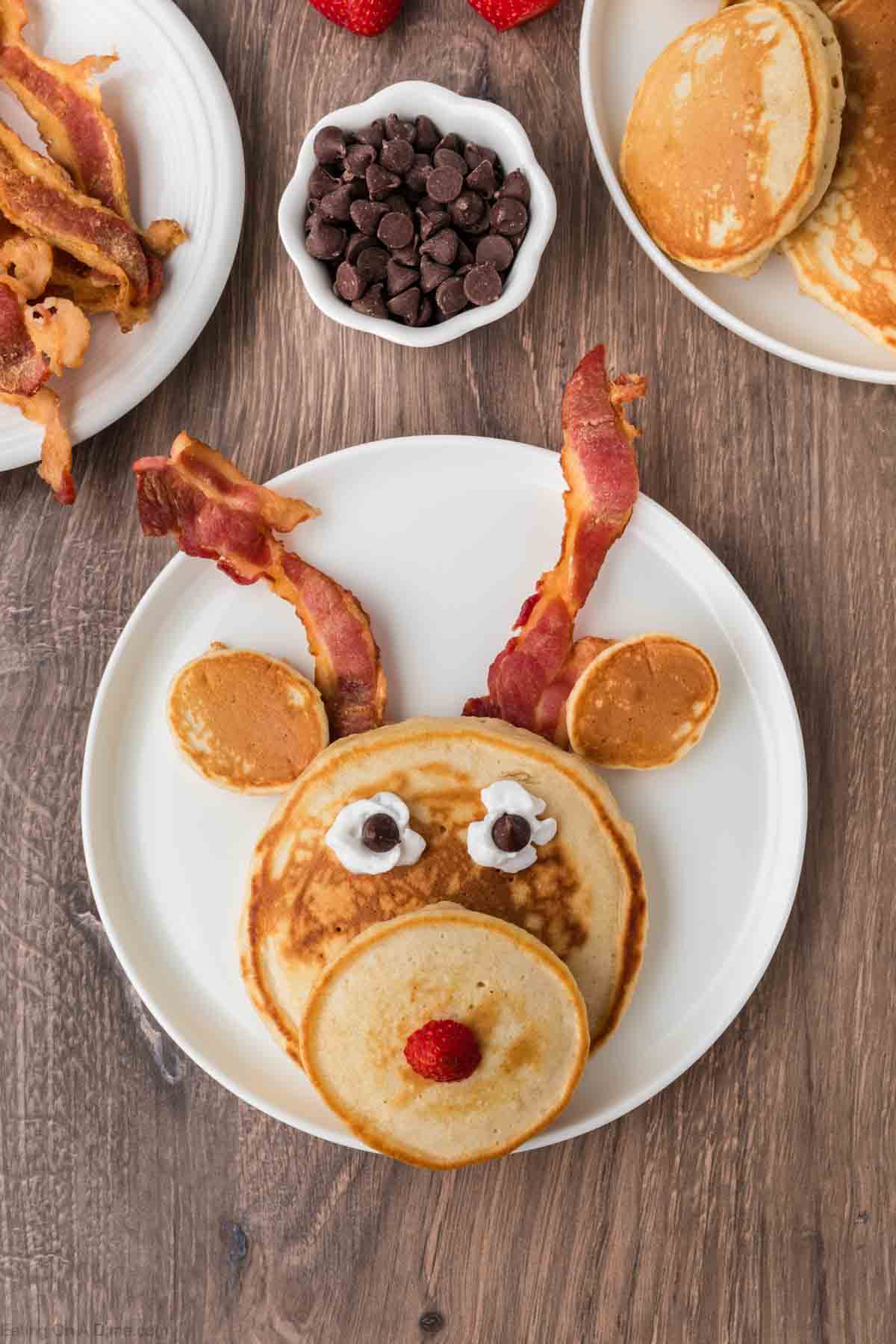 Reindeer face pancakes on a plate topped with whipped cream chocolate chips and raspberries to make the eyes and nose