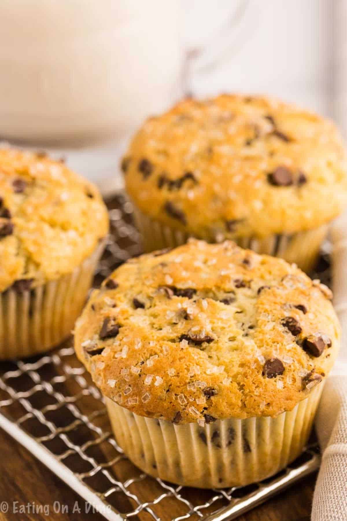 Three golden-brown chocolate chip muffins with sugar crystals on top rest on a wire cooling rack, showcasing the delicious outcome of a favorite Chocolate Chip Muffin recipe. A blurred pitcher is in the background.