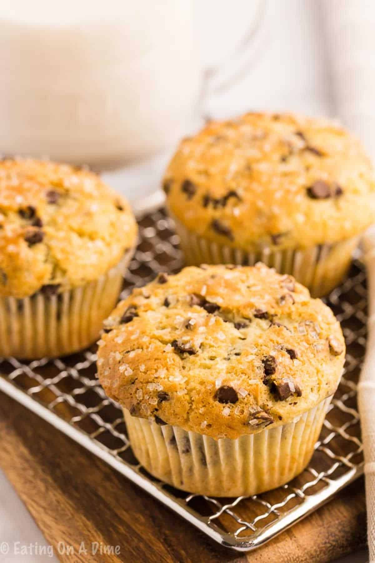 Three chocolate chip muffins rest on a wire rack. Golden brown and dusted with sugar, these treats sit invitingly on a wooden surface, embodying the perfect chocolate chip muffin recipe. A blurred jug adds a touch of rustic charm to the background.