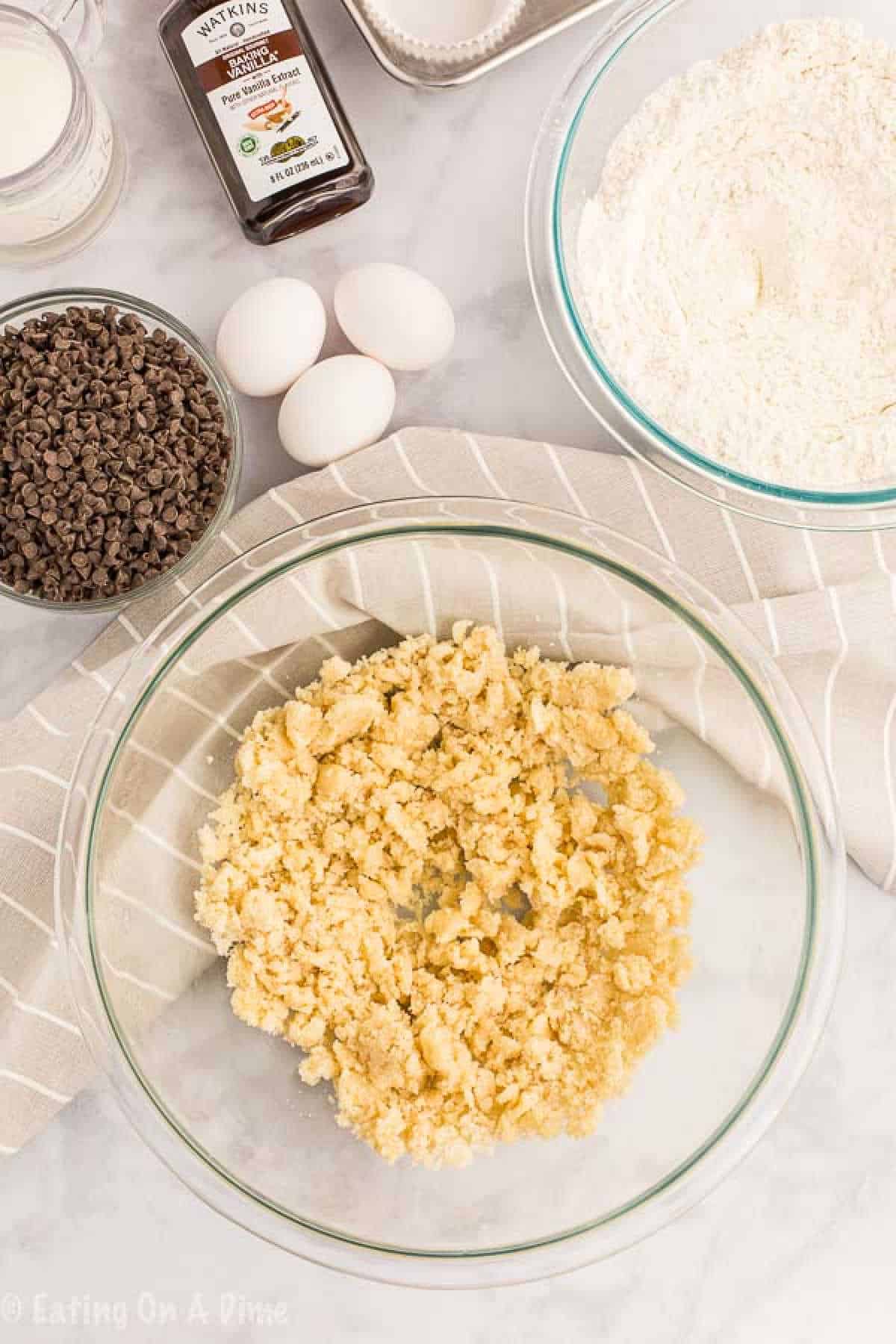 Ingredients for cookie dough on a counter: a glass bowl with crumbly dough, a bowl of flour, three eggs, a bottle of vanilla extract, and a bowl of chocolate chips. These essentials lay the foundation for any sweet treat, whether it's delicious cookies or your new favorite chocolate chip muffin recipe.