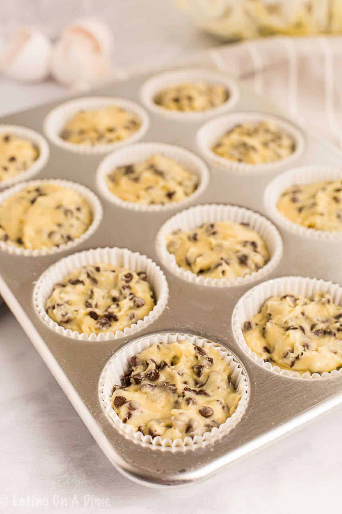 A muffin tray filled with raw chocolate chip muffin batter in white paper liners sits on a light-colored countertop, embodying the delicious promise of a cherished chocolate chip muffin recipe. In the background, a couple of eggs and a bowl add to the scene's cozy anticipation.