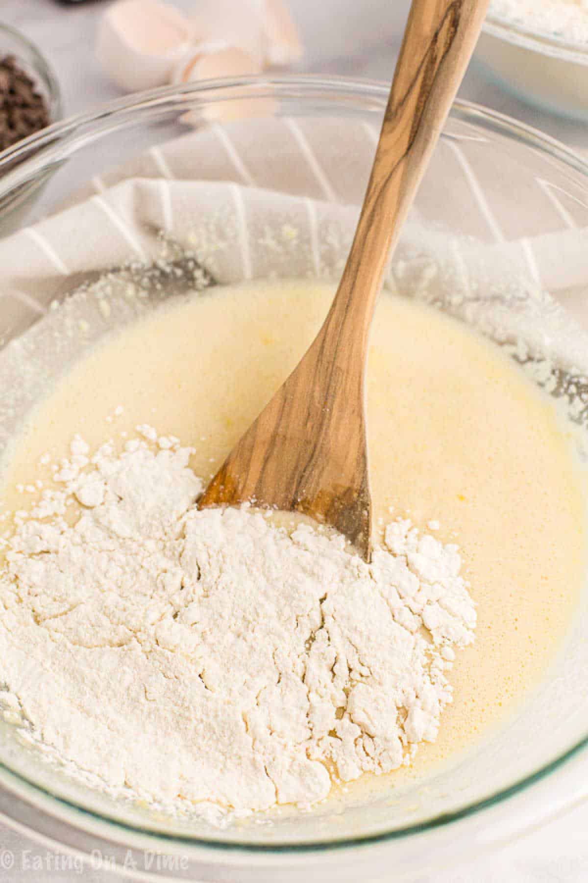 A wooden spoon stirs a chocolate chip muffin mixture of flour and liquid ingredients in a glass bowl. Broken eggshells and a small bowl are softly blurred in the background on a light surface.