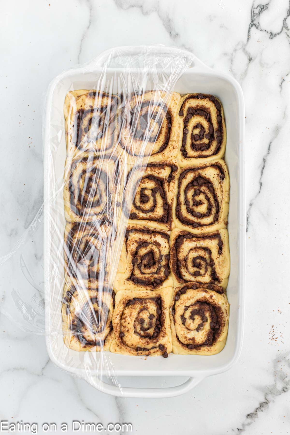 Cinnamon rolls dough in a baking dish topped with plastic wrap