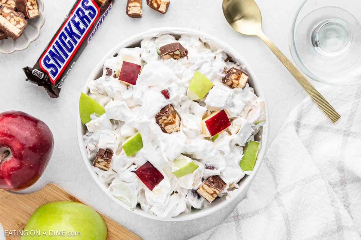 A bowl of 3 Ingredient Snicker Salad with chopped red and green apples, and Snickers pieces mixed in whipped topping. A gold spoon and a Snickers bar lie beside the bowl, while a red apple, a green apple, and an empty glass complete the scene.