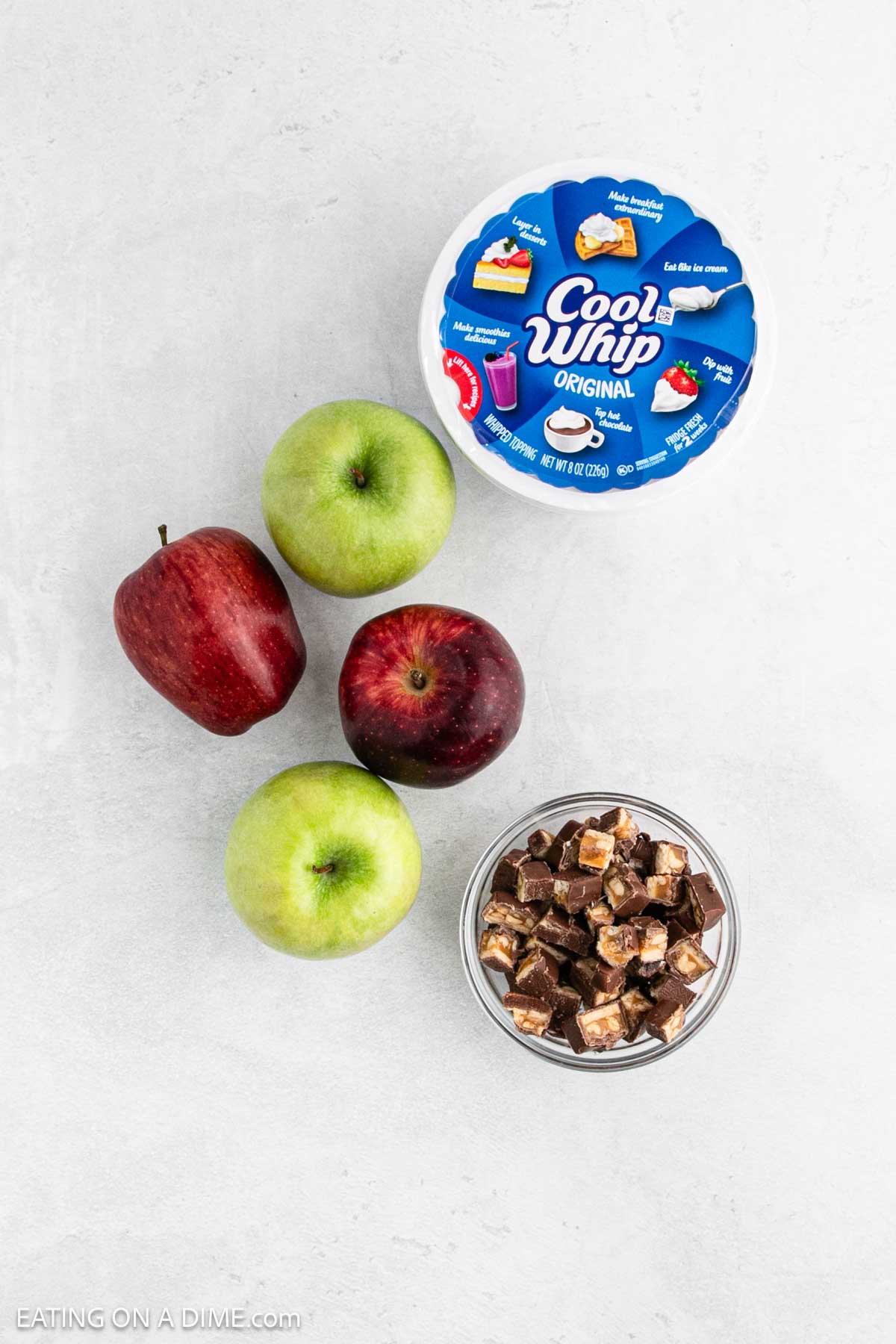 Two red apples and two green apples rest on a white surface beside a bowl of chopped chocolate bars and a tub of Cool Whip, ready to become the perfect 3 Ingredient Snicker Salad.