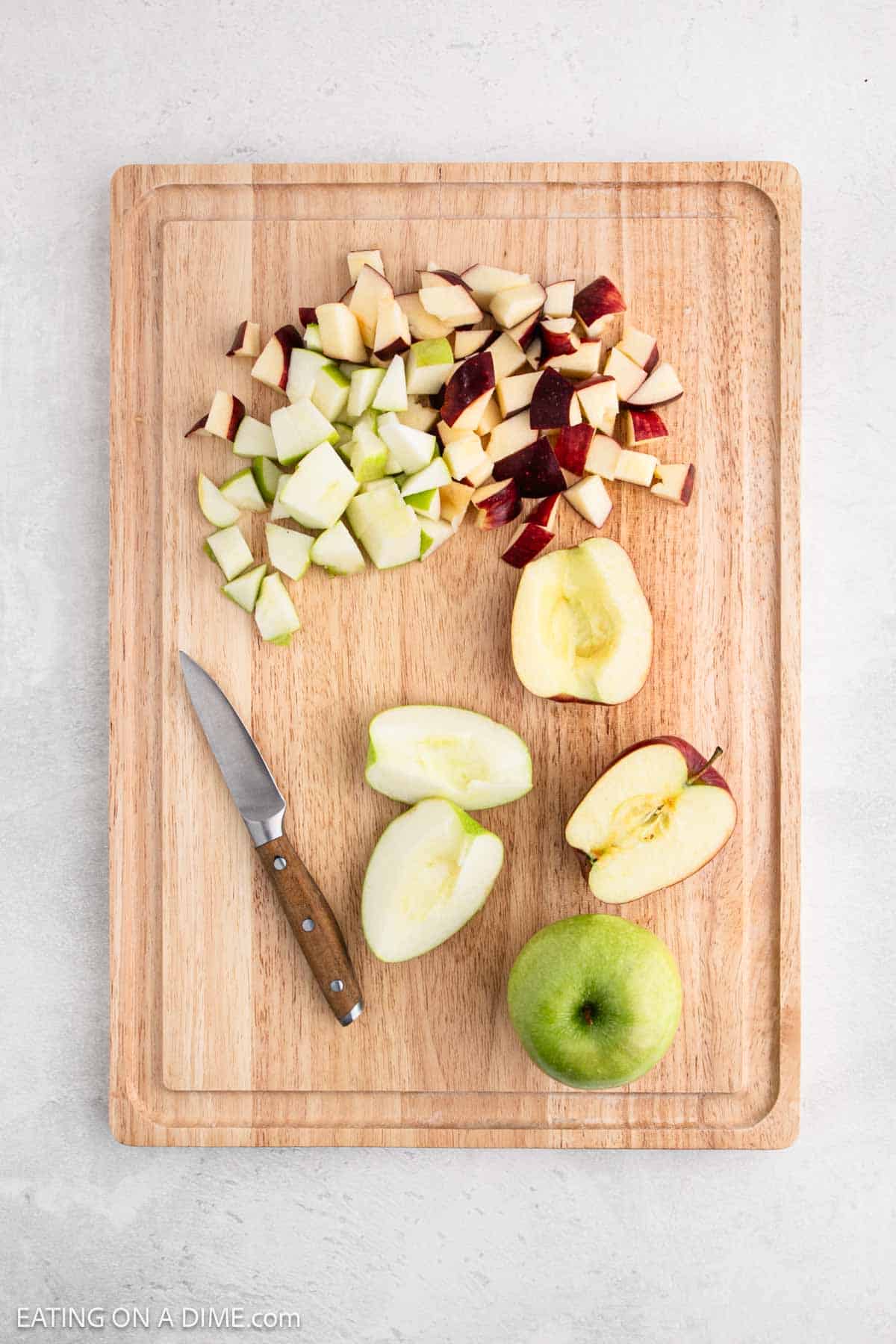 A wooden cutting board showcases the makings of a 3 Ingredient Snicker Salad, with diced red and green apples at the top, a knife on the left, and halved fruit in the center. The apples are captured mid-preparation, hinting at something delicious in every slice.