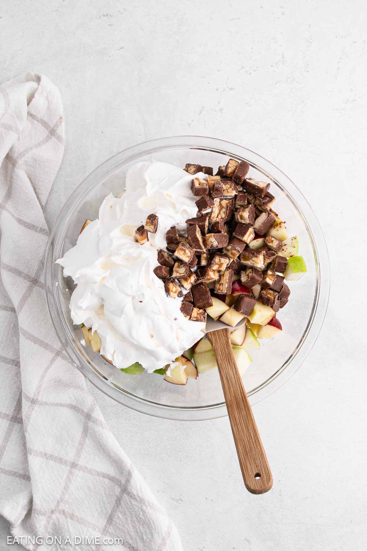 A bowl containing a delightful 3 Ingredient Snicker Salad: chopped apples, whipped cream, and pieces of chopped chocolate bars. A wooden spoon rests in the bowl, and a checkered cloth is placed beside it on a light surface.