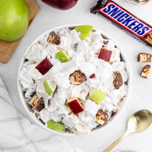 A bowl of 3 Ingredient Snicker Salad awaits, topped with creamy chunks of red and green apples and Snickers candy. A Snickers bar and fresh apples add a tempting backdrop, along with a spoon and napkin nearby.