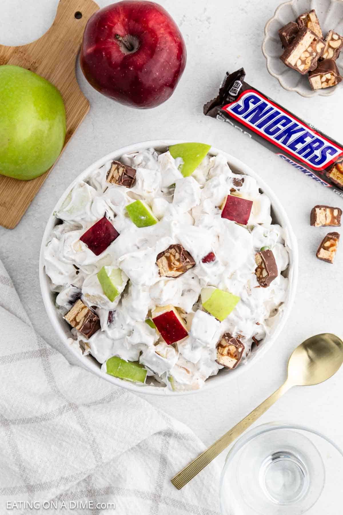 A bowl of 3 Ingredient Snicker Salad combines chopped red and green apples, Snickers pieces, and whipped cream. Nearby, whole apples and a Snickers bar rest beside a gold spoon and a glass of water on a light-colored surface.