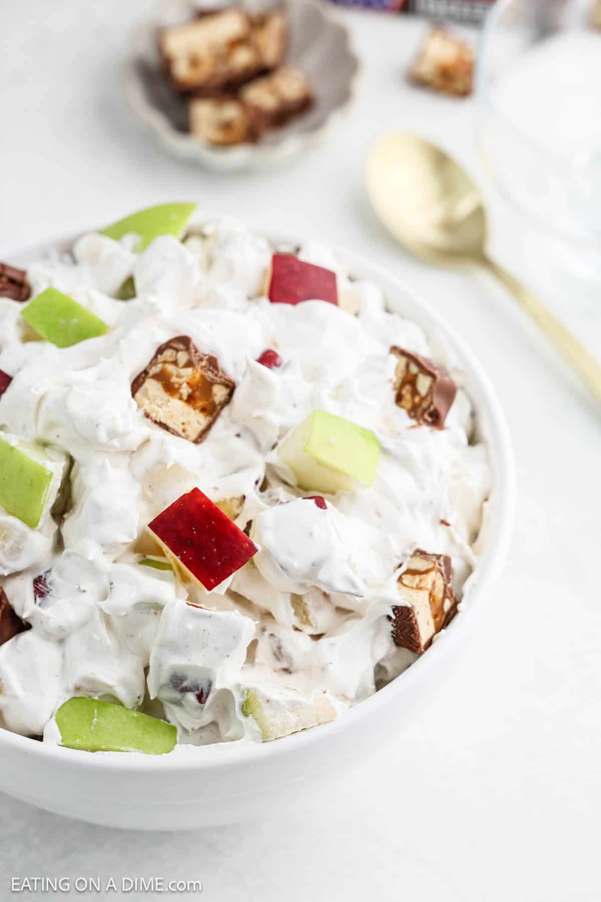 Discover the simplicity of a 3 Ingredient Snicker Salad: a creamy bowl topped with crisp red and green apple pieces and delightful chunks of chocolate-covered nougat. In the background, a gold spoon waits beside an empty glass, ready to enhance your indulgent experience.