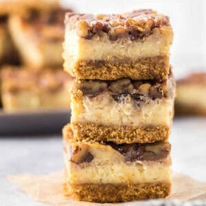 A stack of pecan pie cheesecake bars topped with pecans showcases layers of creamy filling and a graham cracker crust. The bars rest on parchment paper, with more blurred in the background.