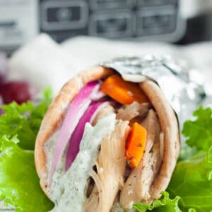 A gyro wrapped in foil featuring succulent slow cooker chicken gyros, red onion, and orange bell peppers, topped with creamy tzatziki sauce. It rests on fresh green lettuce with a blurred kitchen appliance in the background.