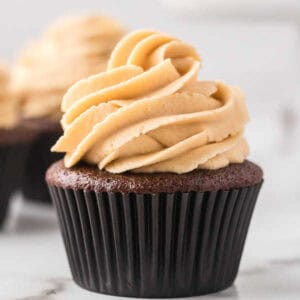 Chocolate peanut butter cupcake with a swirl of creamy, light brown frosting on top, set in a black cupcake liner. The background is blurred, with another similar cupcake partially visible.