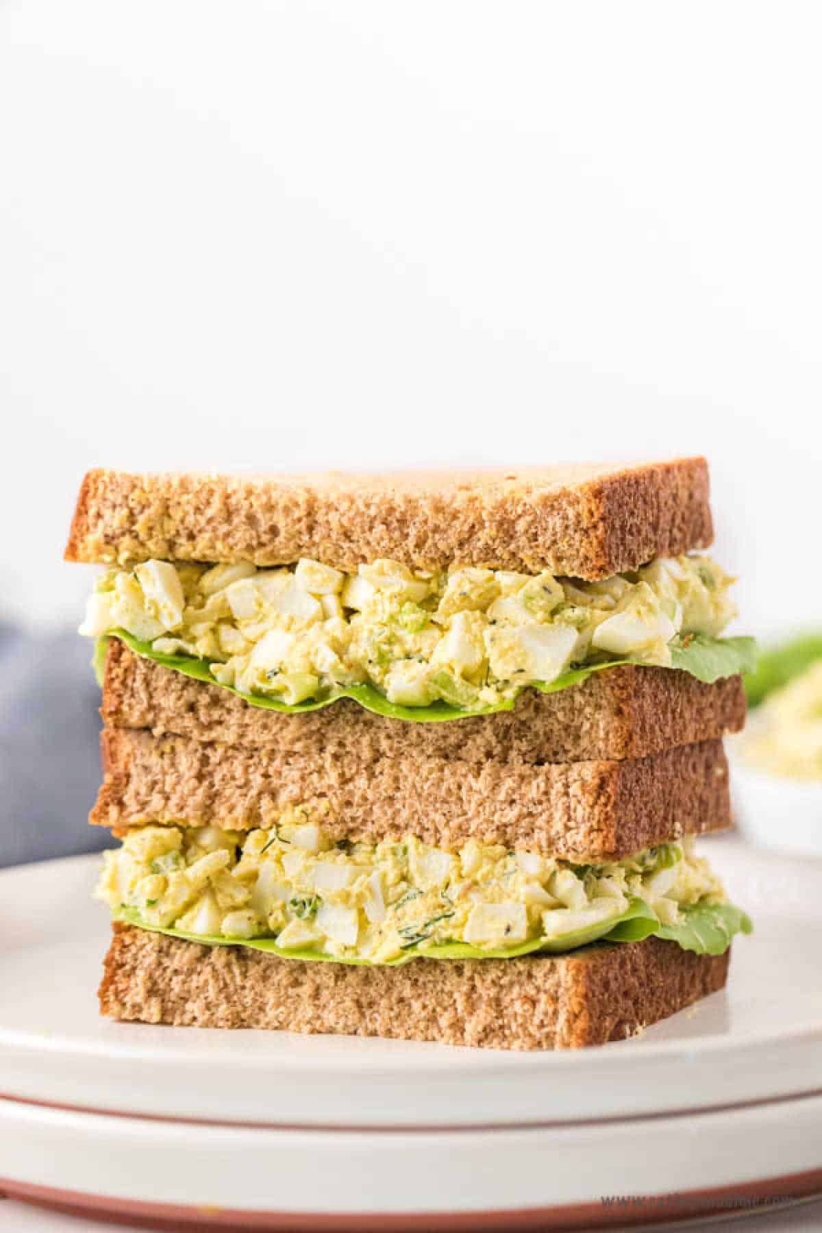 A close-up of two stacked egg salad sandwiches on a plate showcases a delicious egg salad sandwich recipe. The sandwiches feature brown bread filled with a mix of chopped egg salad and crisp lettuce. In the blurred background, a small dish adds to the inviting scene.