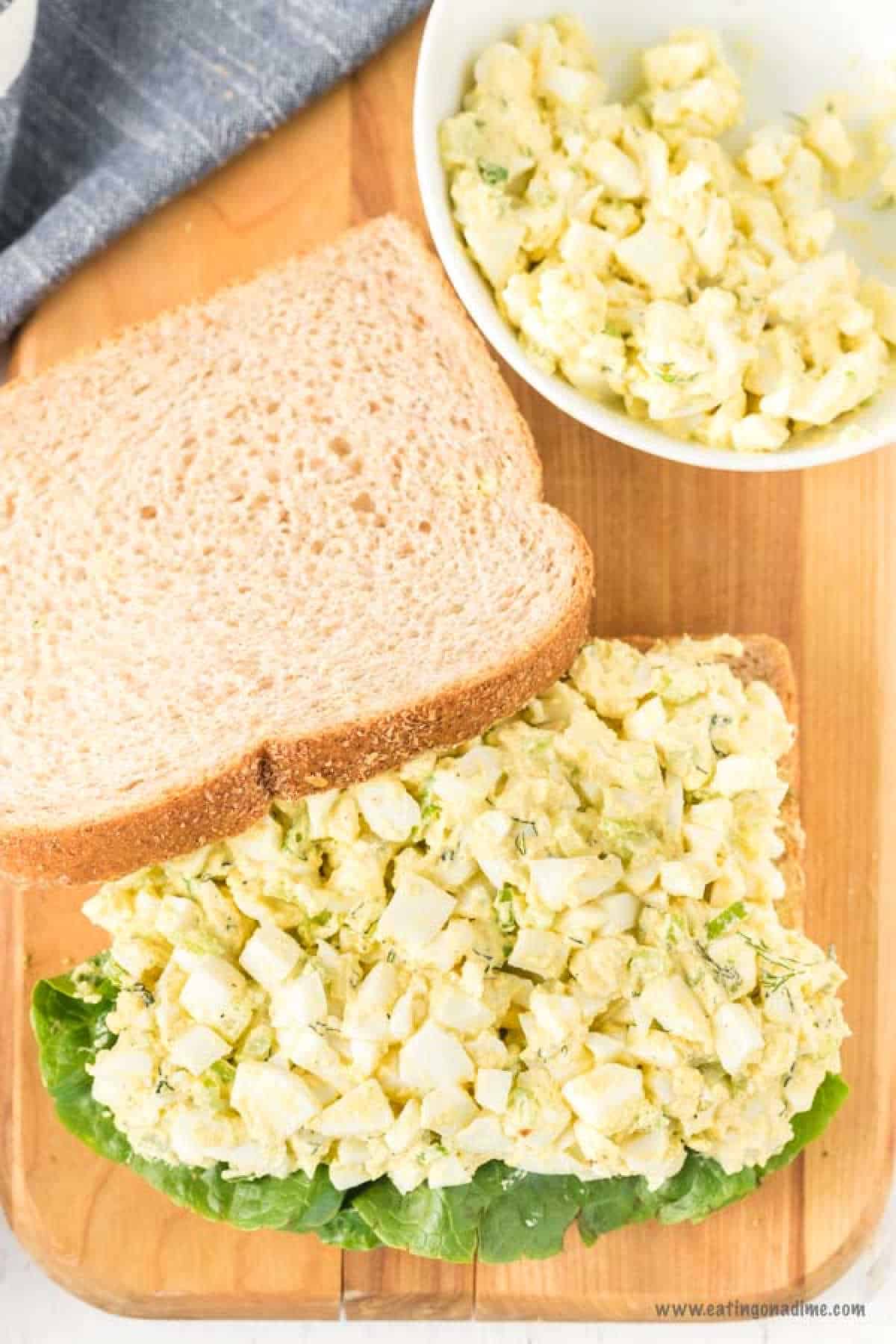 A sandwich on a wooden surface showcases an open slice of bread adorned with egg salad and crisp lettuce, capturing the essence of a classic egg salad sandwich recipe. Another slice of bread lies nearby, ready to complete the creation. In the background, a bowl brims with more egg salad.