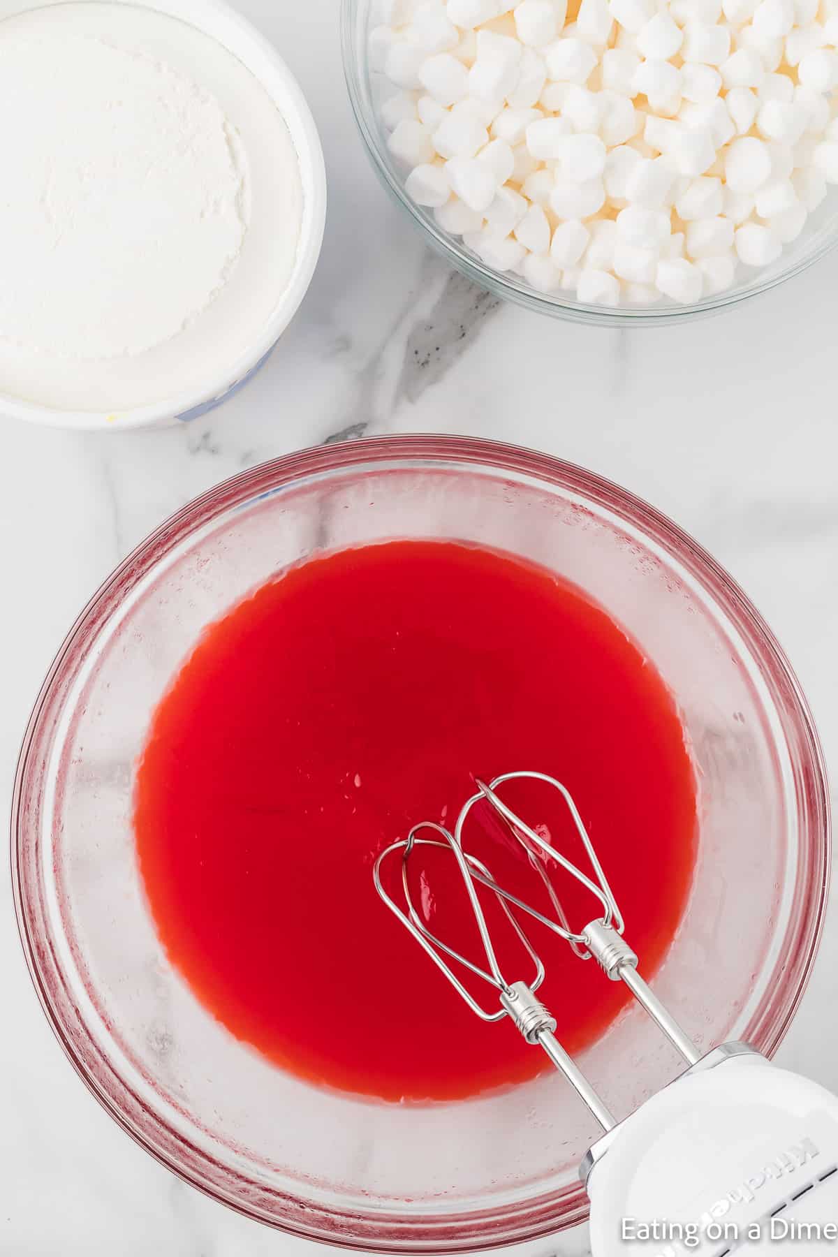 Jello mixture in a bowl with a hand mixer on the side with cool whip and marshmallows on the side