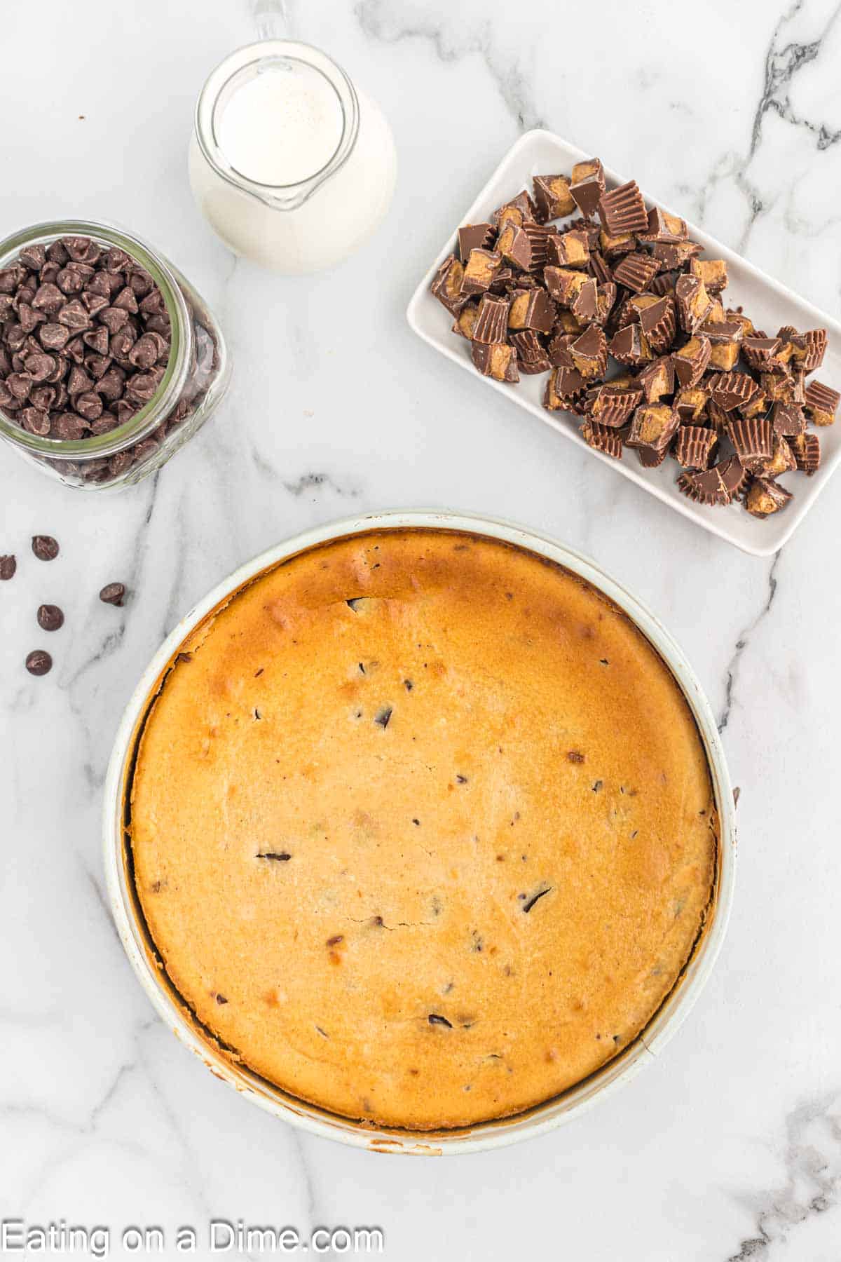 Baked cheesecake in the springform pan with a jar of chocolate chips, heavy cream, and a plate of chopped Reese's Peanut Cups on the side