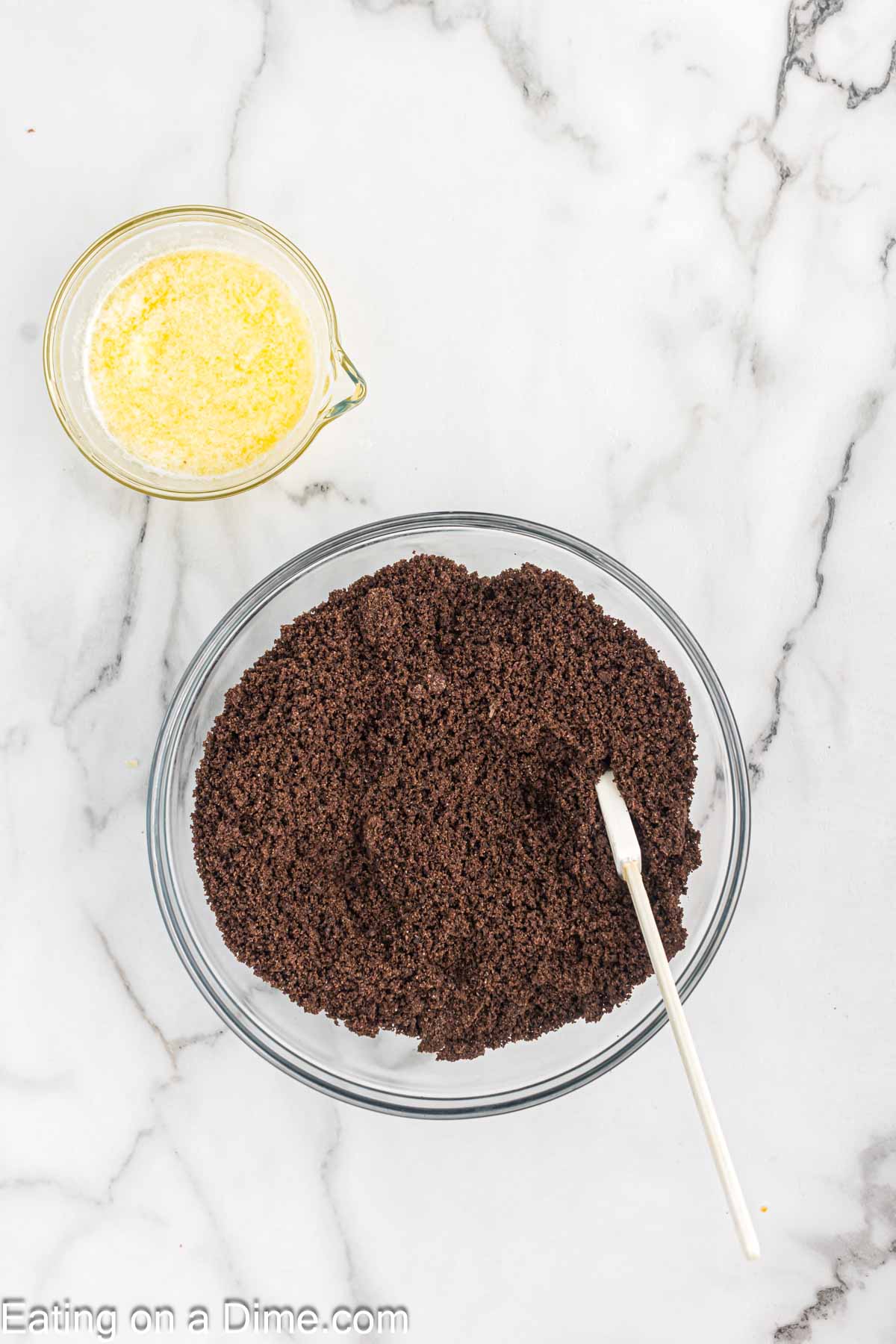 Crushed Oreo Cookies in a bowl with a spatula with a small bowl of melted butter on the side