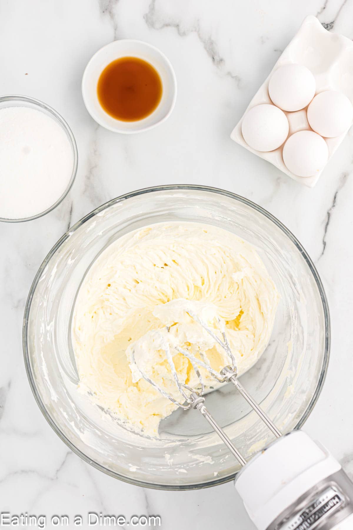 Beating cream cheese in a bowl with a hand mixer with a bowl of heavy cream, vanilla extract and eggs on the side