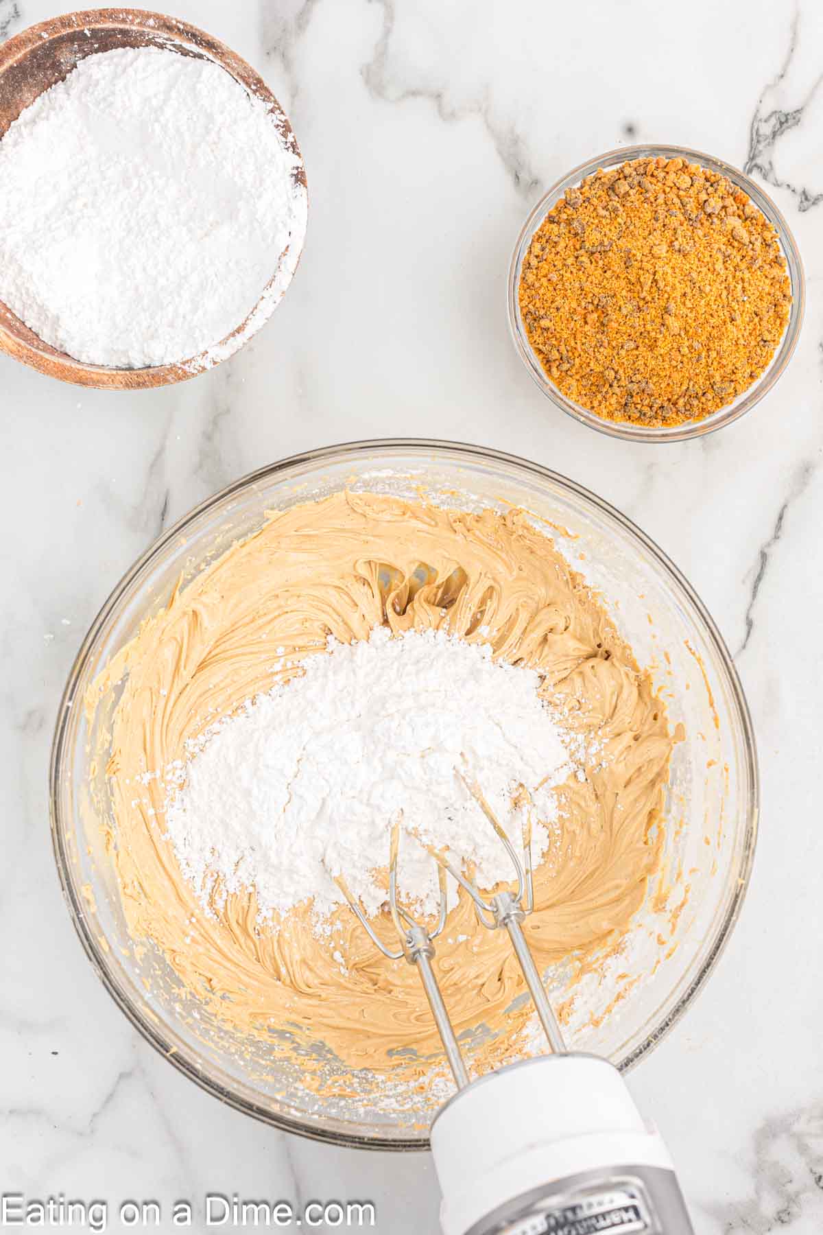 Adding powdered sugar to the peanut butter mixture in a bowl with a hand mixer with a bowl of powdered sugar and butterfinger pieces on the side