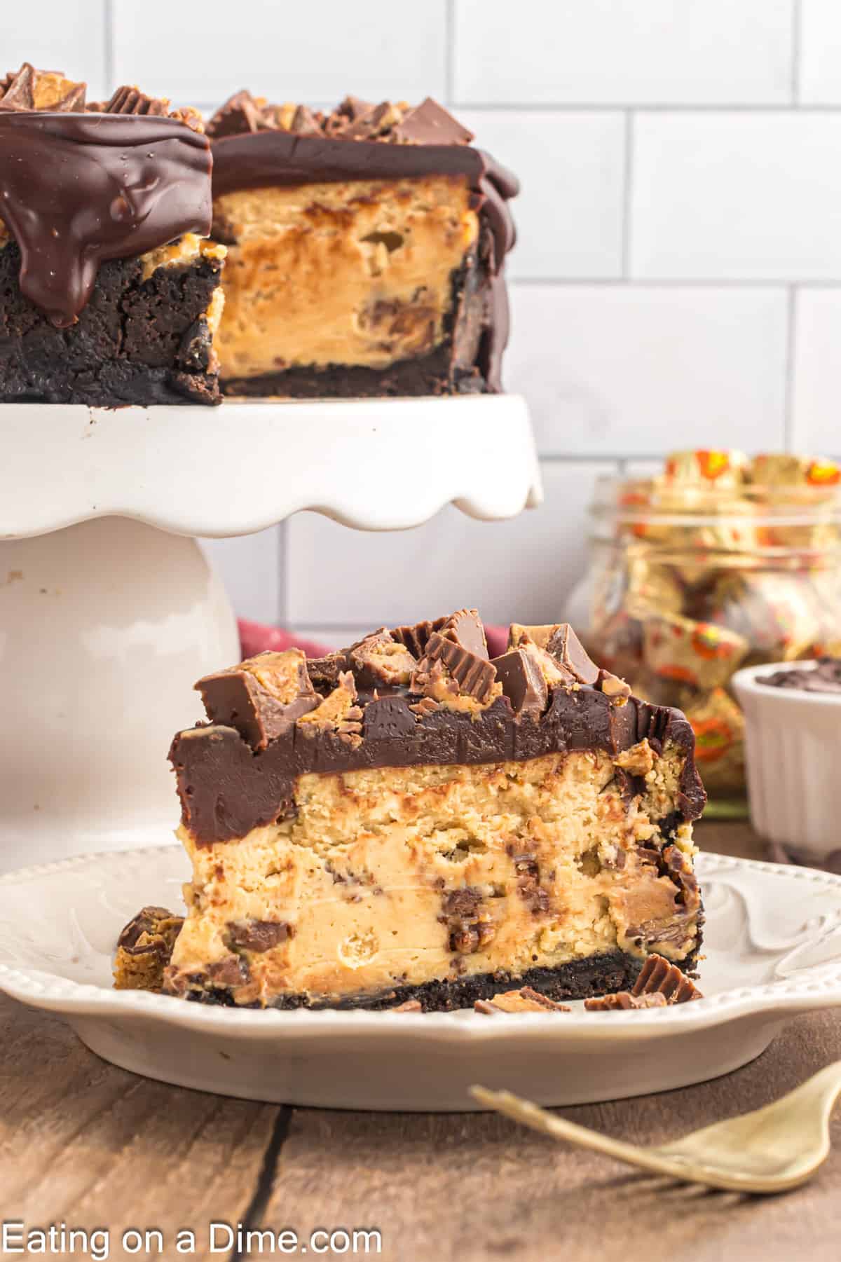 Slice of Reese's Cheesecake on a plate with the Cheesecake on a cake stand with jars of Reese's Peanut Butter Cup in a jar