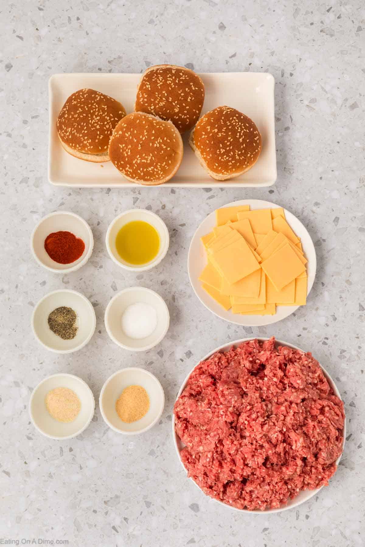 Hamburger buns on a plate, with uncooked ground beef in a bowl with a plate of cheese slices and small bowls of seasoning and oil
