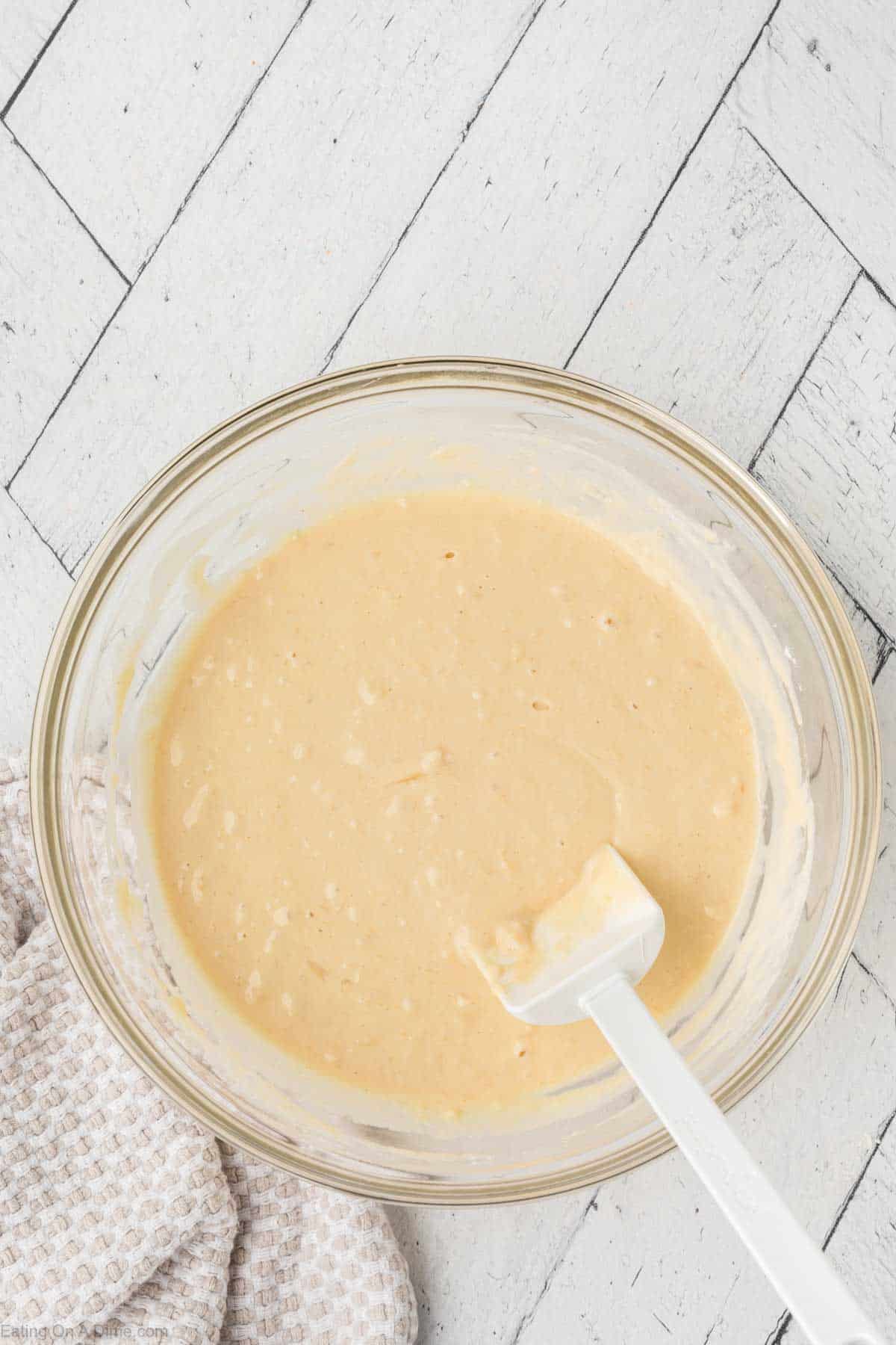 A glass bowl of beige pancake waffle mix sits on a light wood surface. A white spatula rests inside the bowl, ready to stir. A textured cloth is partially visible in the bottom left corner, adding a touch of rustic charm.