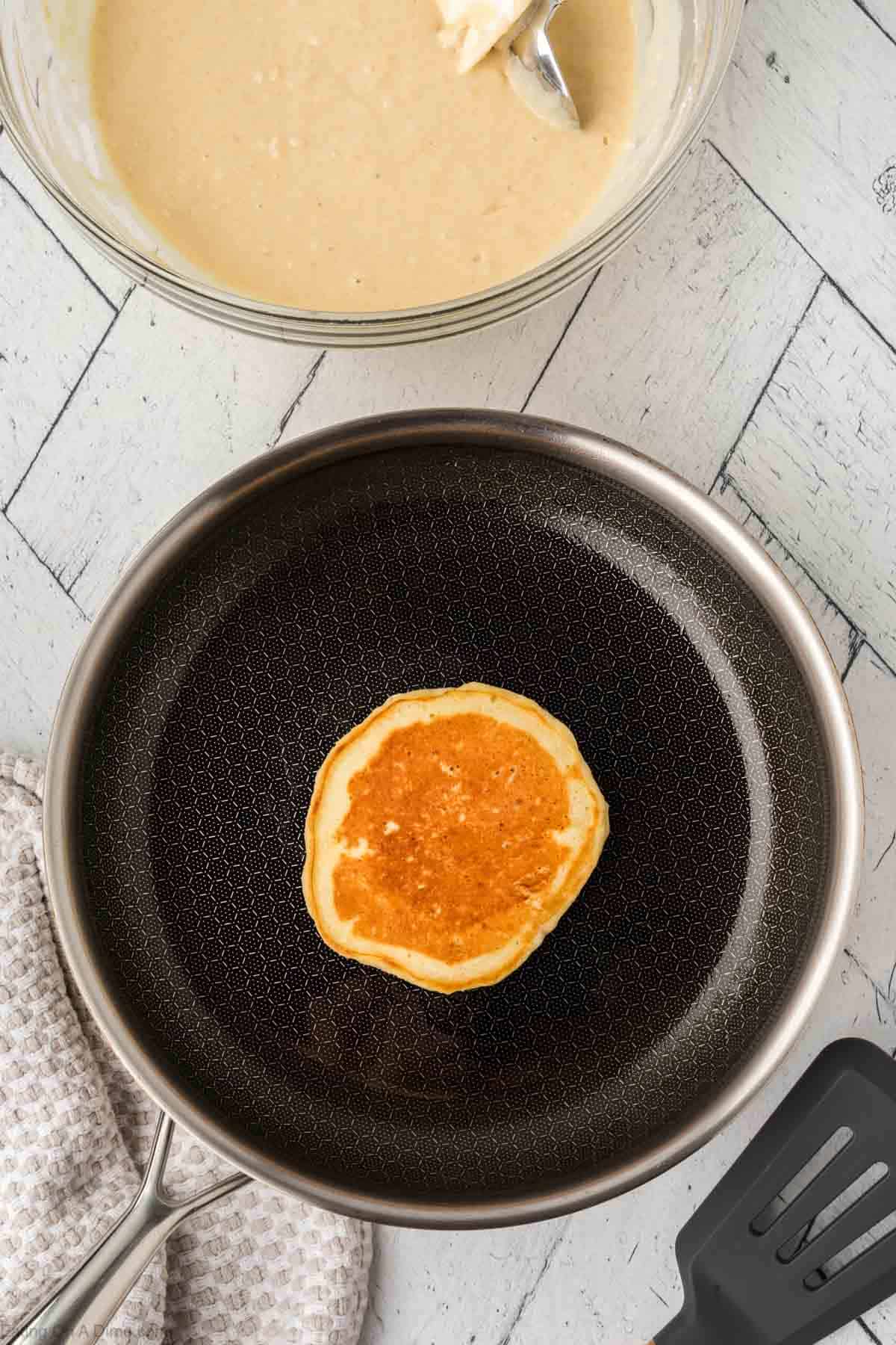 A single pancake cooking in a non-stick frying pan, crafted from a versatile pancake waffle mix. A bowl of batter with a spoon is poised above the pan, while a spatula and cloth rest nearby on the light-colored wooden surface.