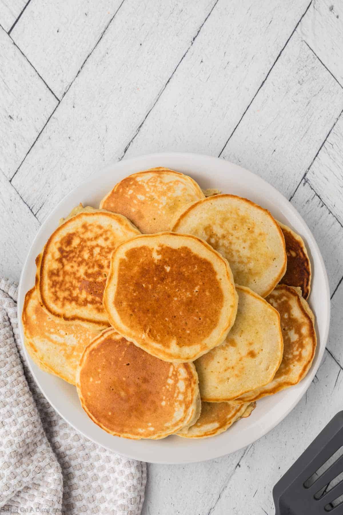 A plate of golden-brown pancakes, crafted with pancake waffle mix, is perfectly stacked on a white table. They're complemented by a checkered cloth and a sleek black spatula.