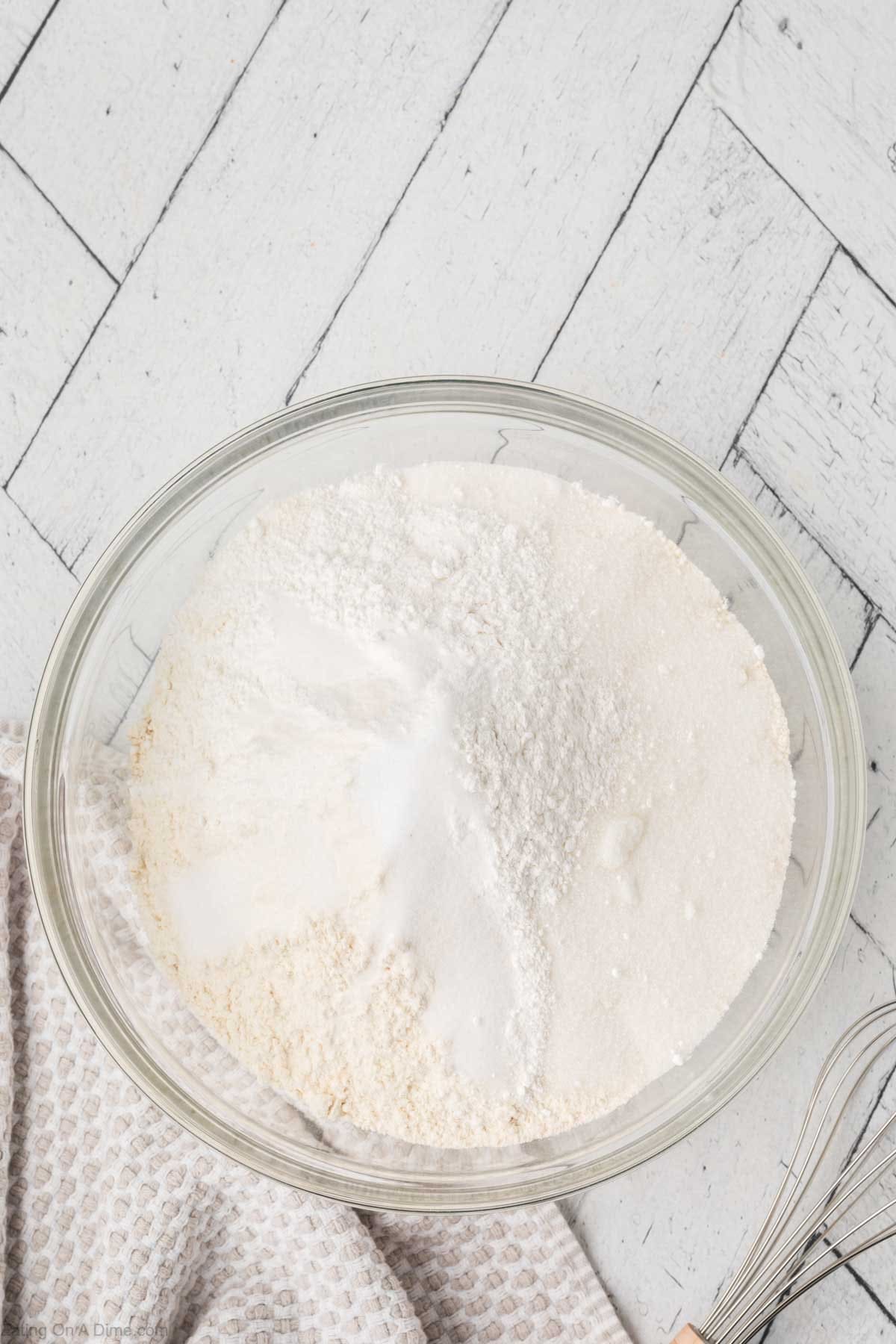 A glass bowl filled with flour and baking ingredients for the perfect pancake sits on a white wooden surface. A whisk and beige patterned cloth are partially visible beside the bowl, ready to mix into these delicious delights.