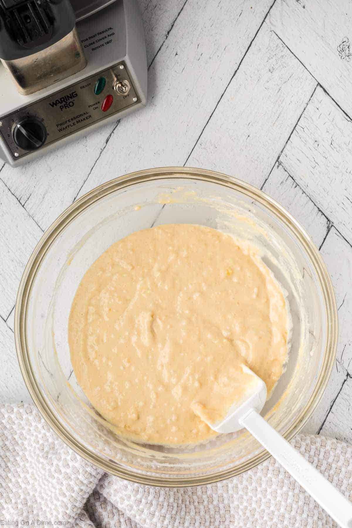 A glass bowl filled with pancake waffle mix sits on a white wooden surface next to a waffle maker. A white spatula rests in the batter, and a checkered cloth is partially visible at the edge of the bowl.