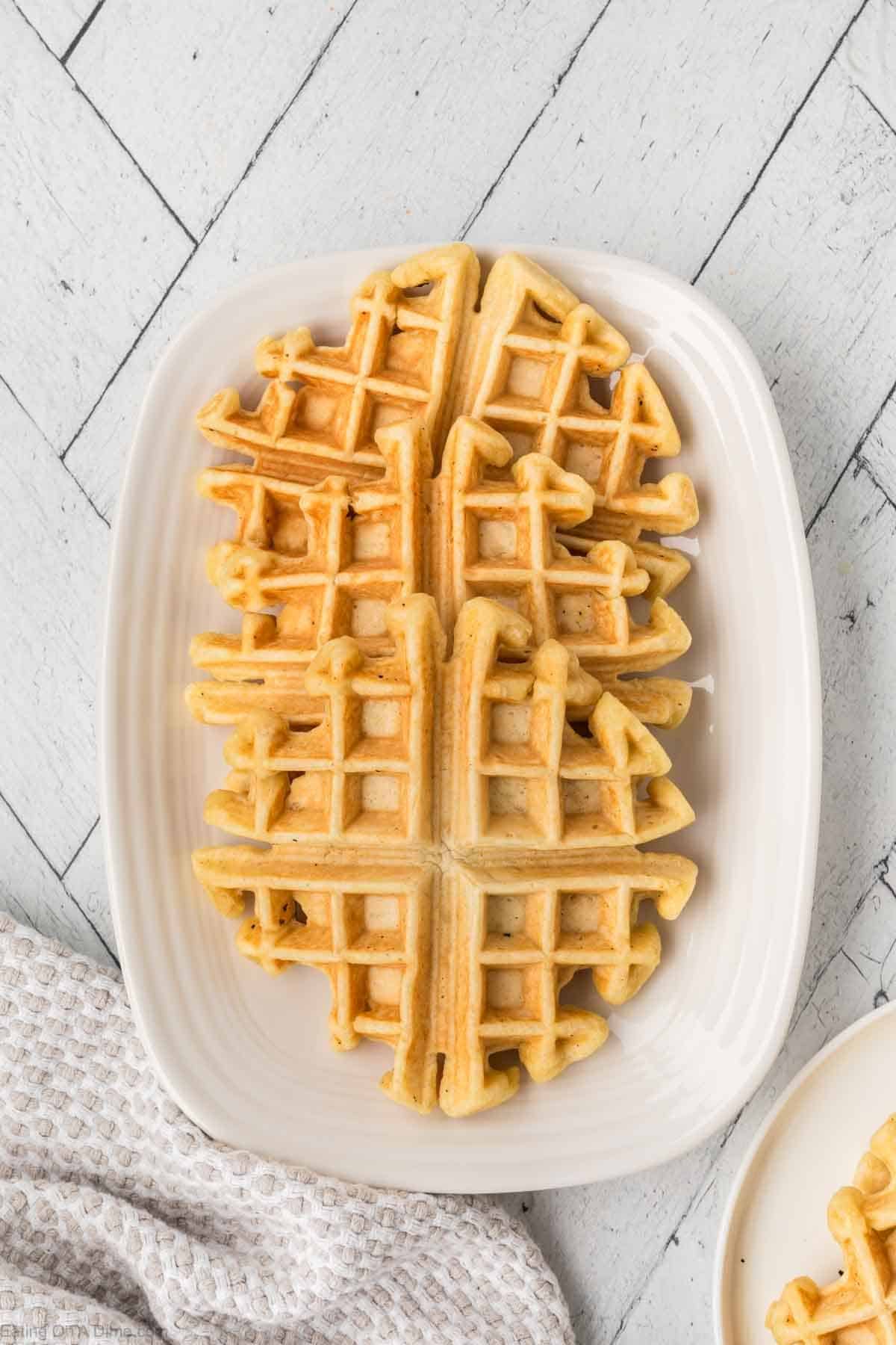 A white rectangular plate showcases four golden-brown waffles made from a delectable pancake waffle mix, arranged in a grid pattern on a light gray, textured surface. A white cloth is partially visible at the bottom left.