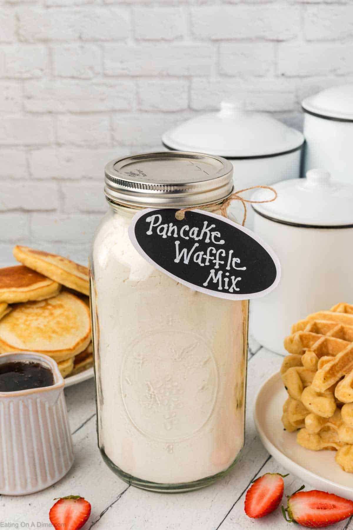 A mason jar labeled "Pancake Waffle Mix" graces the table, surrounded by fluffy pancakes and crispy waffles. A small dish of syrup and fresh strawberry slices add a touch of sweetness, while white kitchen canisters stand against a rustic brick wall.