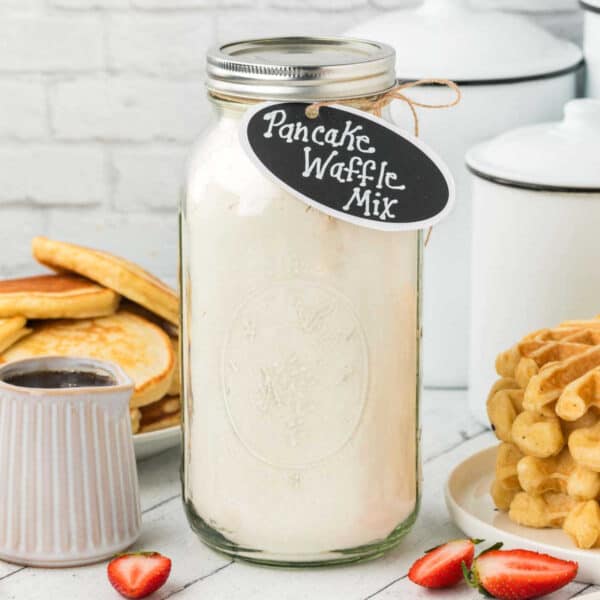 A jar labeled "Pancake Waffle Mix" sits enticingly on a table, encircled by fluffy pancakes, crispy waffles, a small jug of syrup, and vibrant sliced strawberries. The inviting scene is framed by a light brick wall and accompanied by sleek white storage containers.