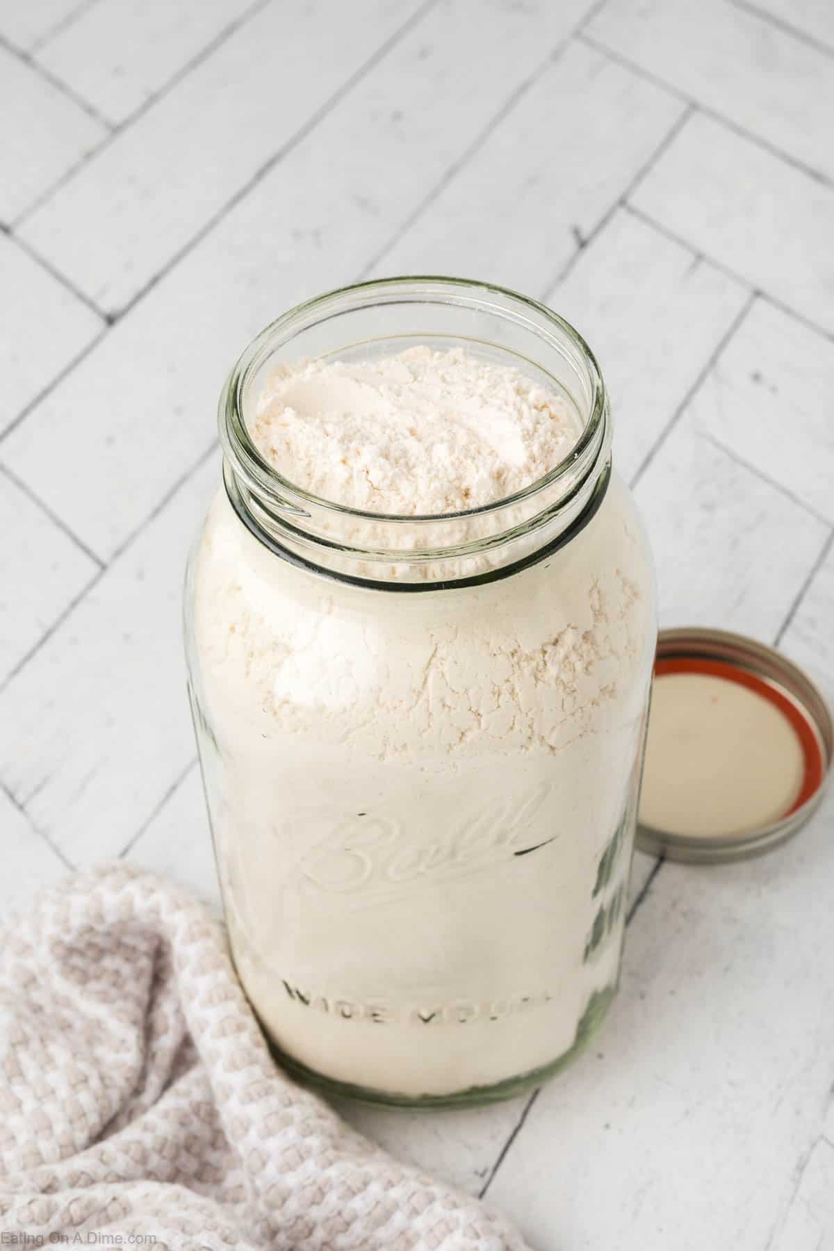 A large glass jar filled with white pancake mix, with a screw-on metal lid resting beside it. The jar is placed on a light-colored, herringbone-patterned surface, next to a folded white and gray textured cloth.