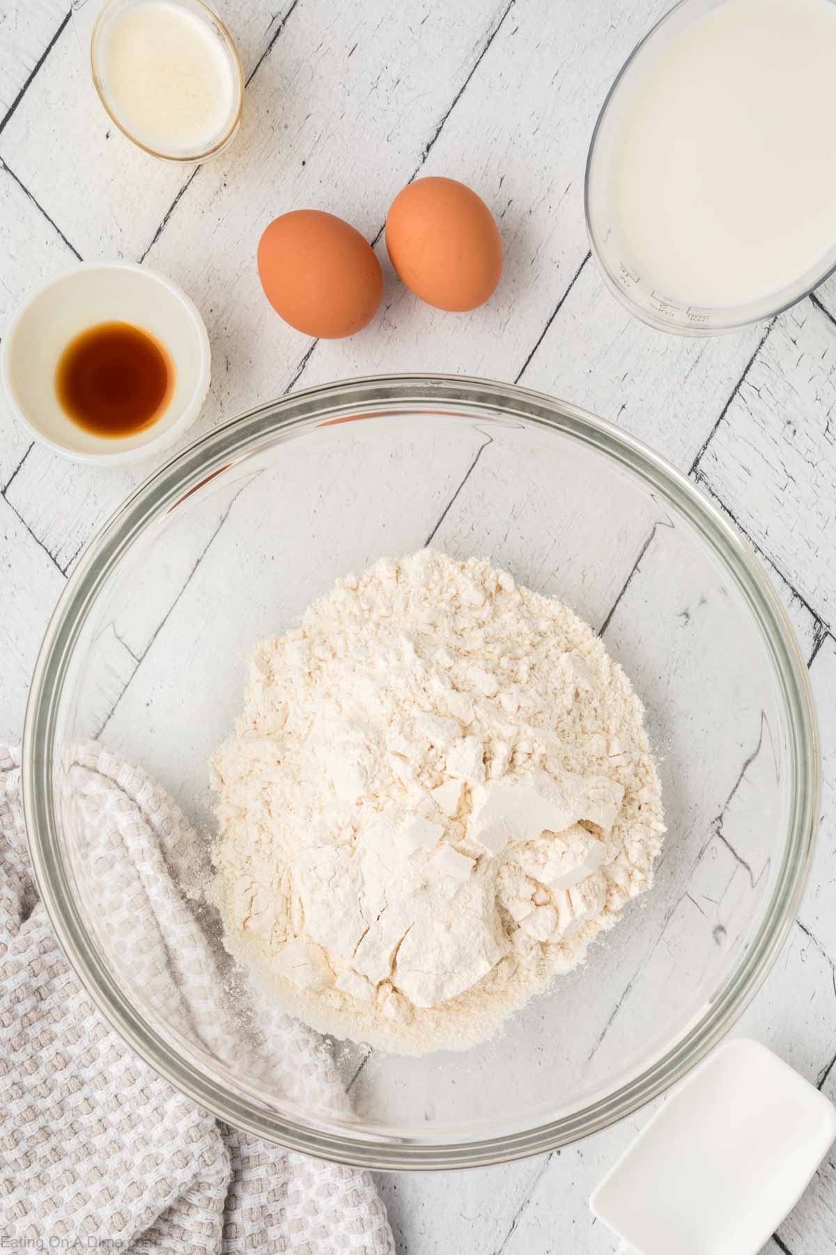 A bowl of flour on a white wooden surface, surrounded by two eggs, a pitcher of milk, a small bowl of vanilla extract, and a bowl with a liquid—perfect beginnings for pancake waffle mix. A textured cloth is partially visible on the side.