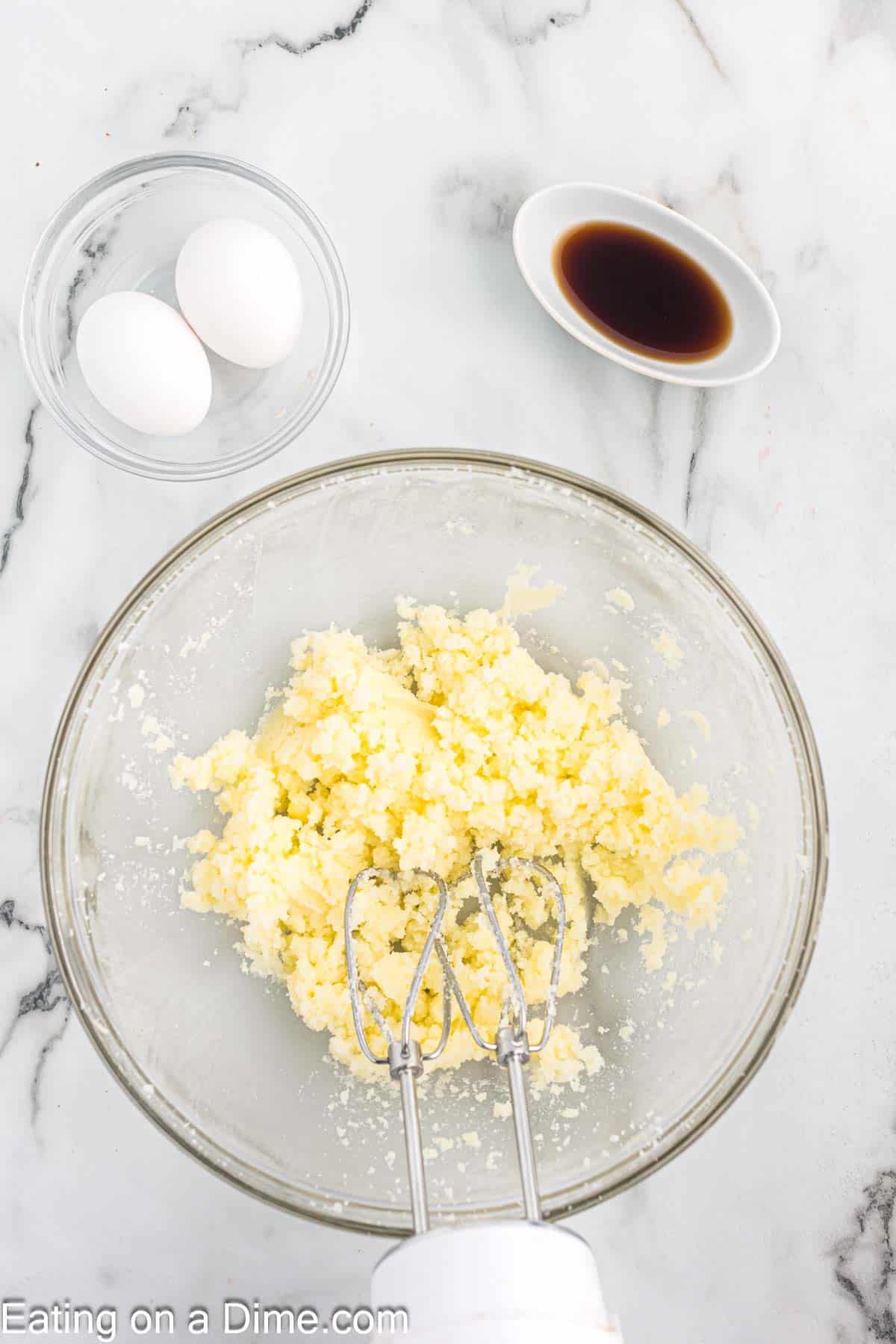 Beating butter and sugar in a bowl with a hand mixer with a small bowl of two eggs and vanilla extract