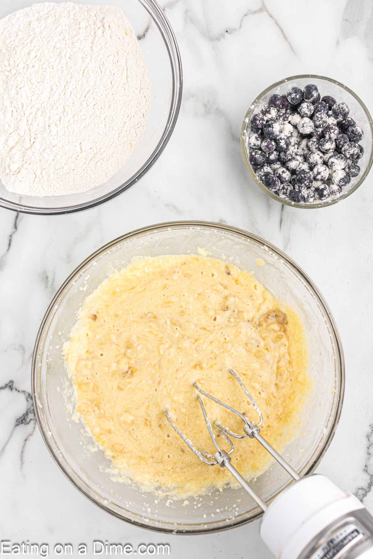 Combining the mashed bananas in the bowl with the butter mixture with a bowl of flour and flour coated blueberries in a bowl