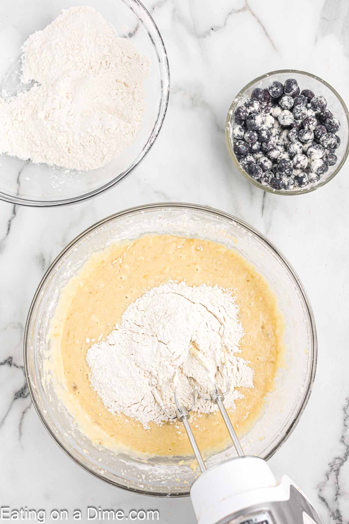 Adding the dry ingredients to the wet ingredients in a bowl with a hand mixer with a bowl of flour and a bowl of flour coated blueberries