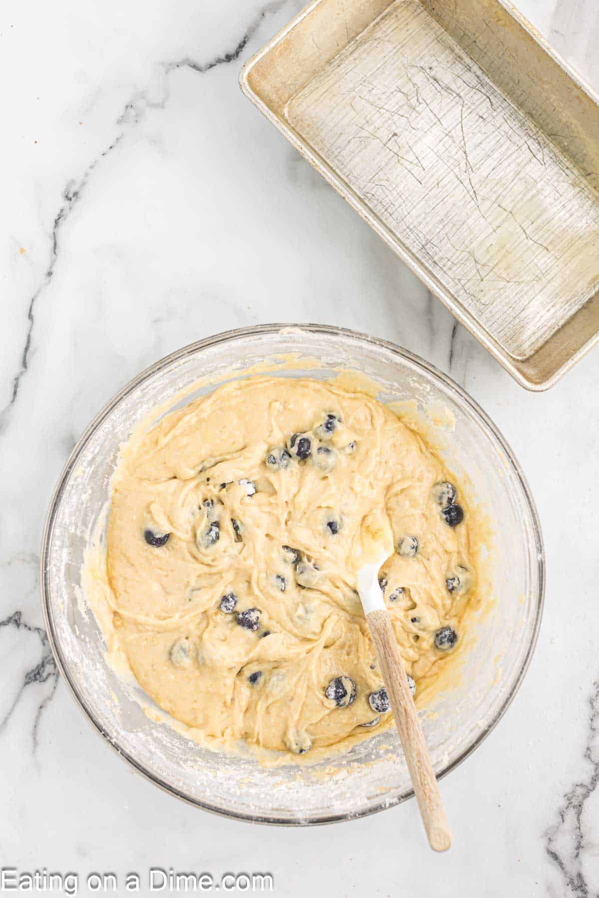 Mixing the blueberries in the bowl of the banana batter with a loaf pan on the side