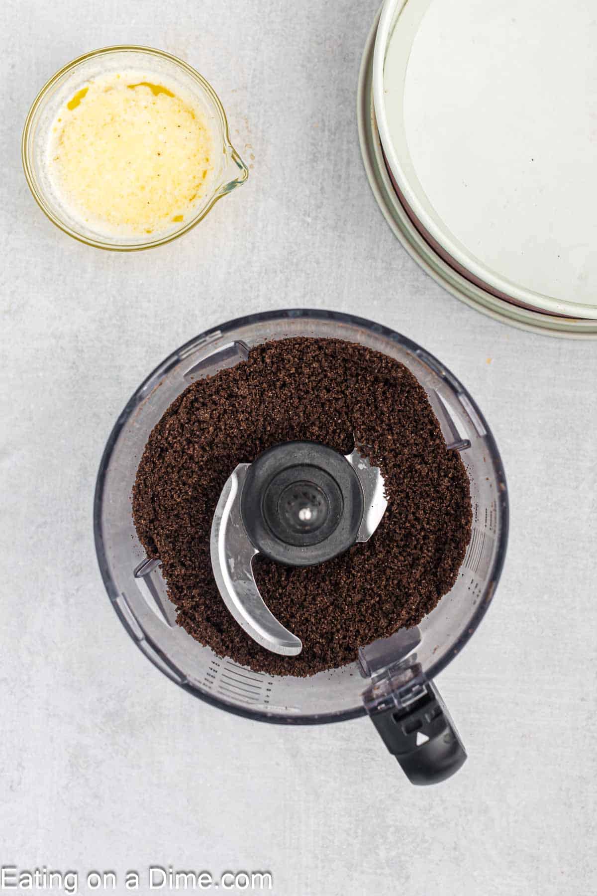 Crushing Oreo Cookies in a food processor with a side of melted butter and a pie pan