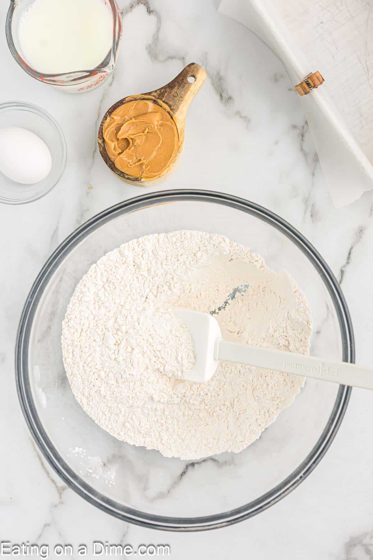 Combining dry ingredients in a bowl with a scoop of peanut butter on a measuring cup with a egg in a bowl