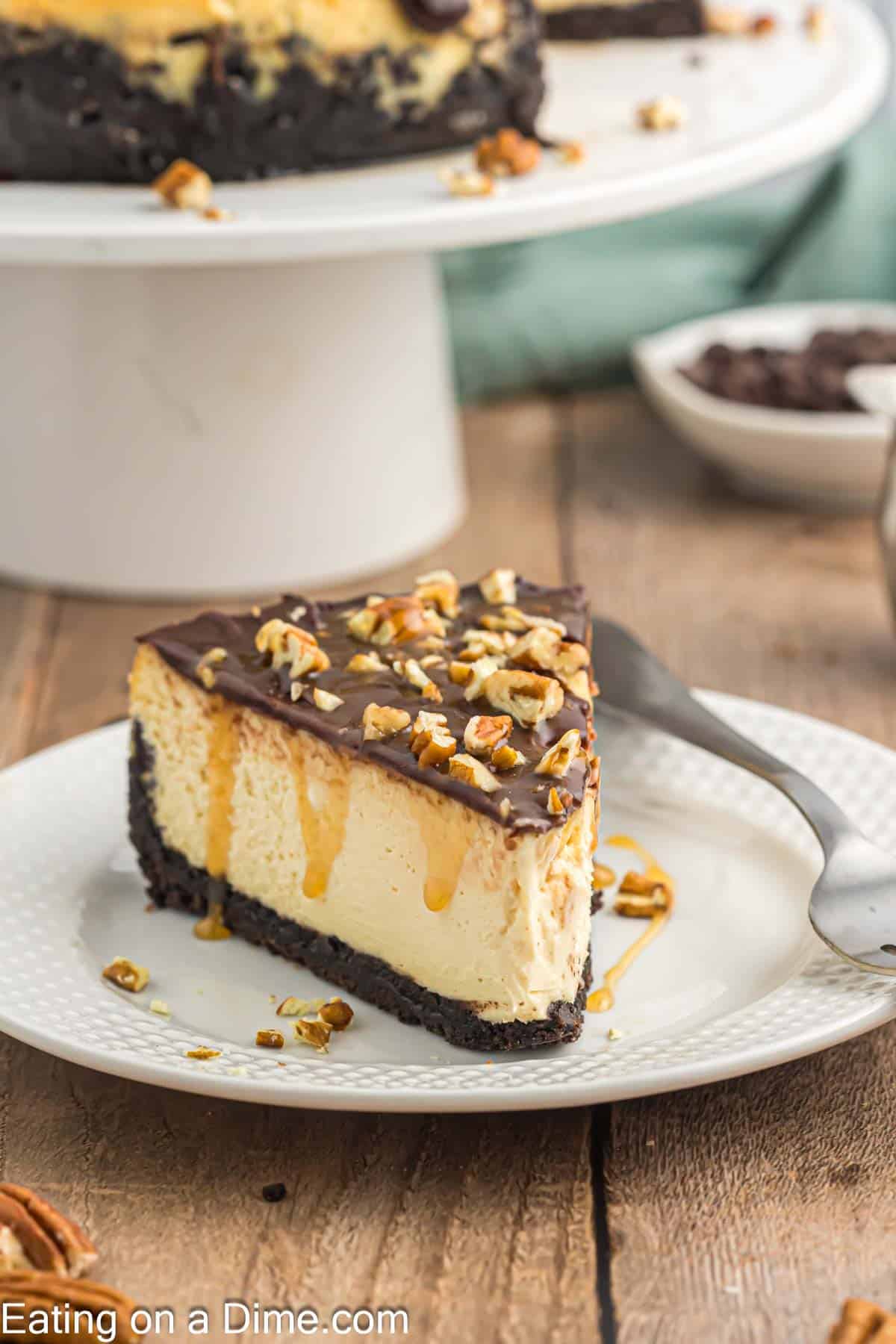 Slice of Turtle Cheesecake on a plate with Turtle Cheesecake on a cake stand