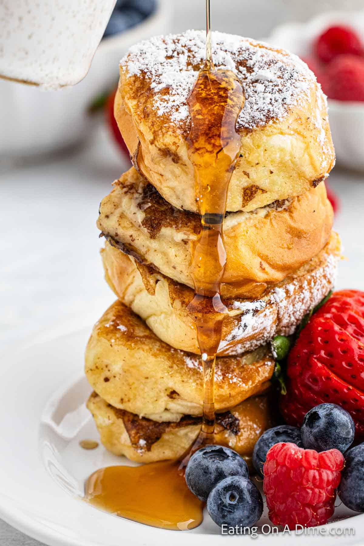 Hawaiian Rolls French Toast stacked on a plate topped with maple syrup and a side of fresh fruit