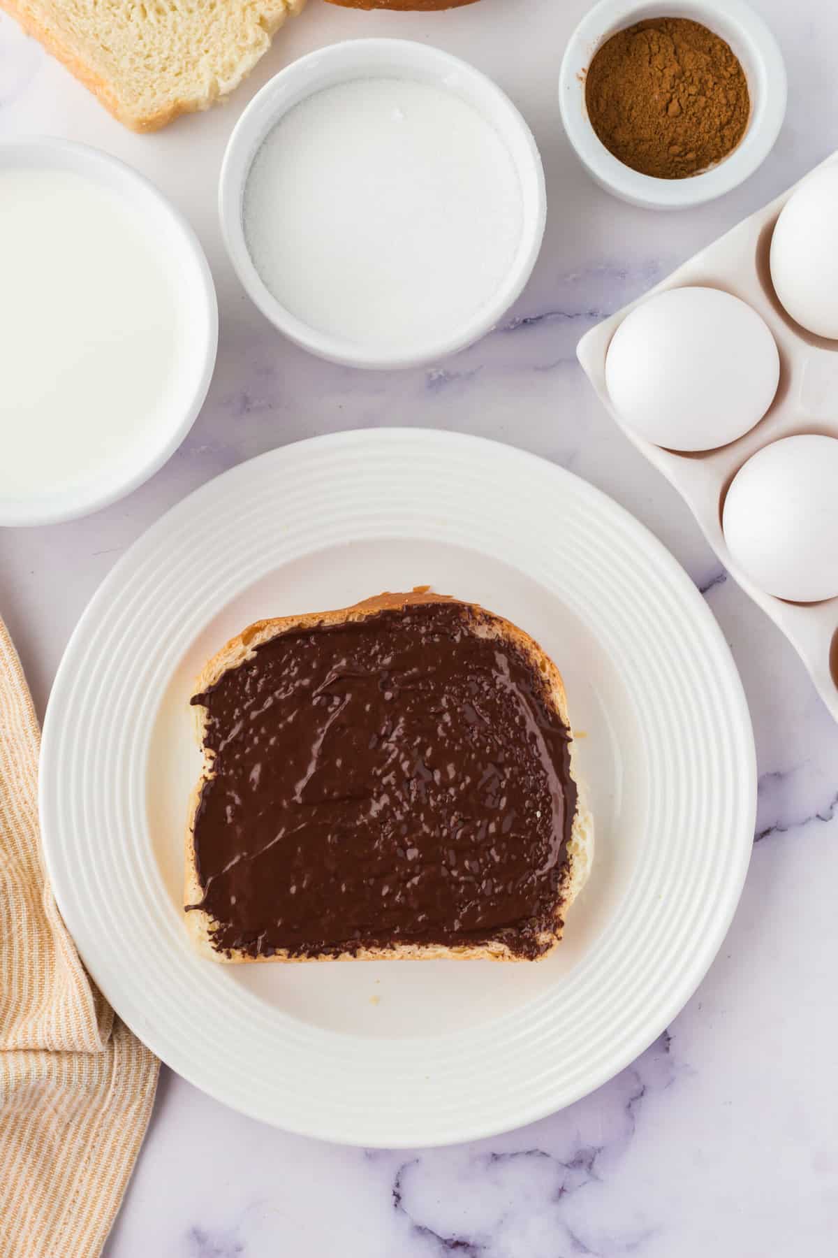 Nutella spread over the top of a piece of bread on a plate with a bowl of milk, sugar and cinnamon the side with a carton of eggs
