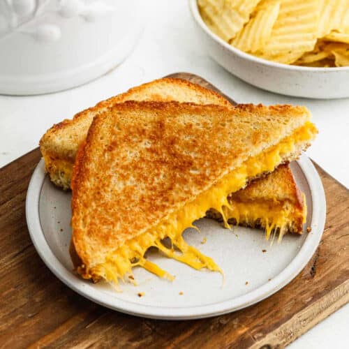 A perfectly grilled cheese sandwich, sliced in half with gooey cheese spilling out, sits invitingly on a white plate atop a wooden cutting board. In the background, a bowl of ridge-cut potato chips awaits your indulgence.